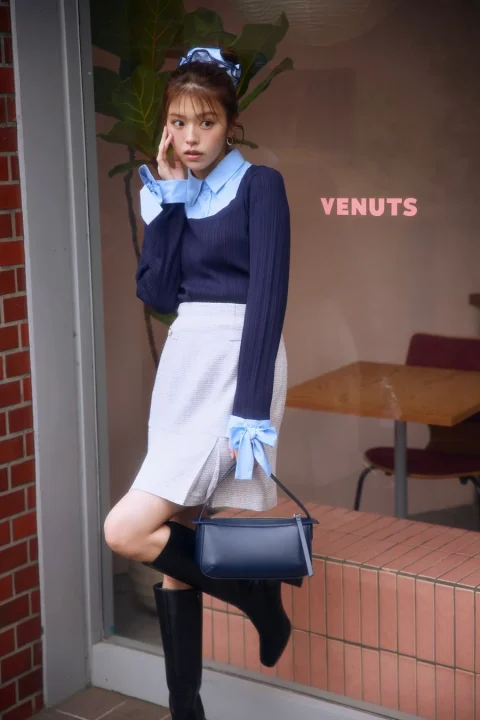 ネイビーのニットと水色のシャツを重ね着し、白いスカートと黒いブーツを合わせた女性が、屋外でポーズをとっています。ヘアアクセサリーと袖のリボンがアクセントになっています。