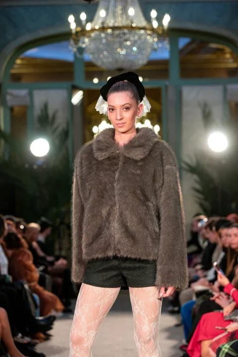 茶色のフェイクファージャケット、黒のショートパンツ、白いレースタイツを着用し、頭には黒いヘッドピースをつけたモデル