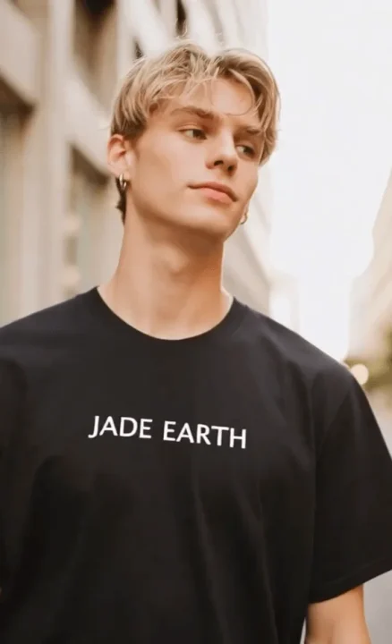 金髪の若い男性が黒いTシャツを着て屋外に立っており、Tシャツには「JADE EARTH」と書かれています。彼は横を向いていて、イヤリングをしています。