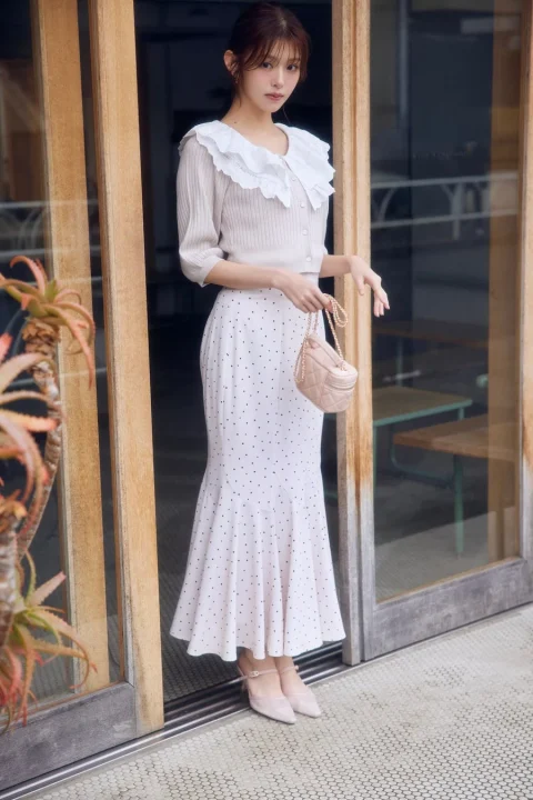 エレガントなフリル襟のニットトップスとドット柄のマーメイドスカートを着用した若い女性が、木製ドアの前に立っています。ピンクのミニバッグとヒールを合わせた、上品でフェミニンな全身コーディネートです。