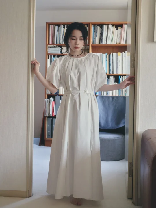 白いロングワンピースを着た若い女性が、本棚のある部屋の入り口に立っている