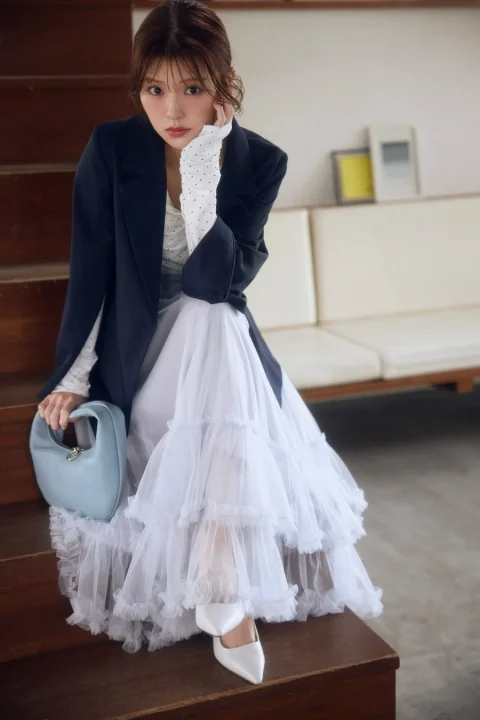 階段に座る女性のファッションポートレート。黒いジャケットに白いチュールスカート、ドット柄のインナーを合わせた大人フェミニンなコーディネート。水色のバッグと白いパンプスがアクセントになっている。