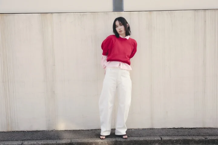 短い黒髪の女性が、赤いセーターとピンクのシャツ、白いワイドパンツを着用