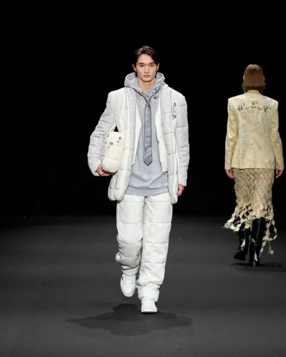 ファッションショーのランウェイを歩くモデルの画像。男性モデルは白いキルティングのセットアップにグレーのパーカーとネクタイを着用し、女性モデルは背中に文字の入ったジャケットを着ています。