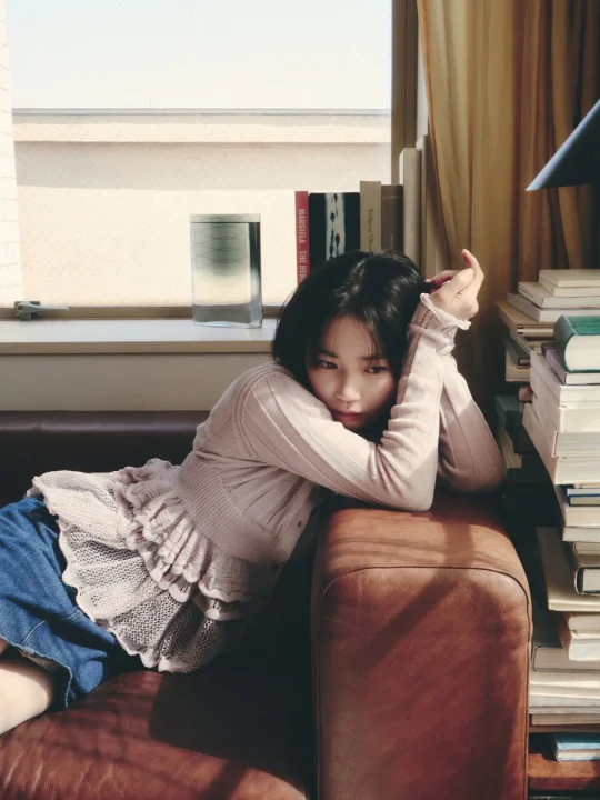 茶色のソファに横たわる若い女性が、窓から差し込む自然光の中でリラックスした表情