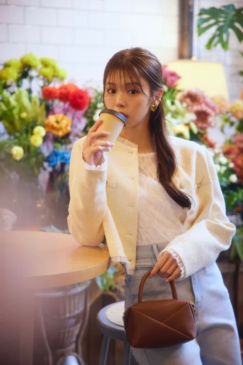 花が飾られた明るい空間で、コーヒーカップを口元に運ぶ女性。白いトップスとクリーム色のジャケット、淡い色のジーンズを着用し、ブラウンのハンドバッグを持っている。おしゃれで落ち着いた雰囲気のポートレート。