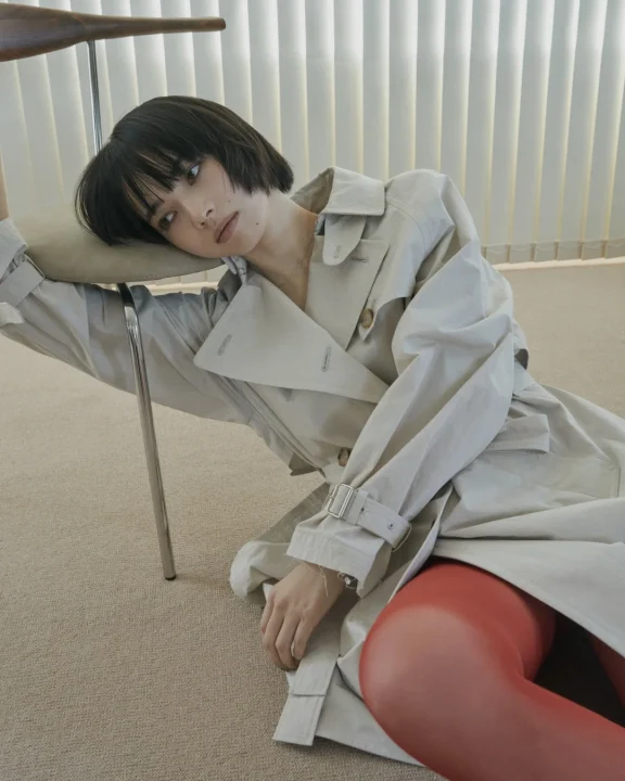 トレンチコートと赤いタイツを着用し椅子に寄りかかる小松菜奈さん