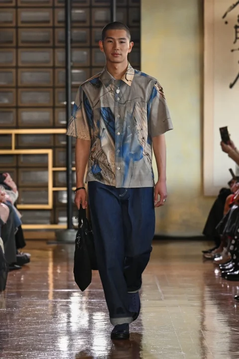 抽象的な青とベージュの柄の半袖シャツとワイドなデニムパンツを着用した若い男性モデル。