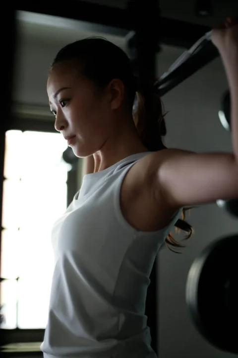 トレーニングウェアを着た若い女性が、集中した表情でジムで運動している様子です。バーベルのような器具を使い、健康的なライフスタイルを思わせる一枚です。