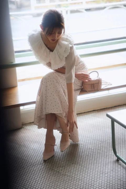 白いフリルブラウスとドット柄スカートを着た女性が、明るい窓辺に座っています。淡いピンクのハイヒールを履き、小さなハンドバッグが隣に置かれ、優雅でフェミニンな雰囲気を醸し出しています。