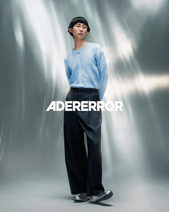ADERERRORのモデルがライトブルーのセーターとダークなワイドパンツ、個性的なシルバーの靴を着用している画像