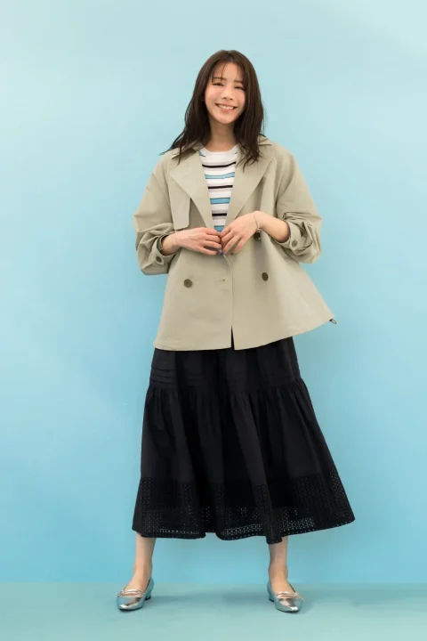 水色の背景の前に立つ、笑顔の女性の全身像です。ベージュのショート丈ジャケットにストライプのトップス、裾にデザインのある黒いロングスカートとシルバーのフラットシューズを合わせた、明るくカジュアルなコーディネートです。