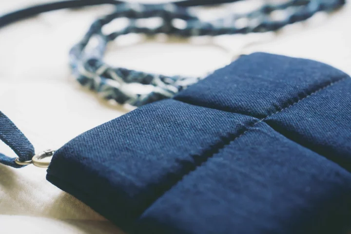 濃い青色のデニム調の生地で作られた、キルティング加工が施されたポーチまたは小型バッグのクローズアップです。ストラップと金属製のリングが見え、手作りの温かみが感じられる布製品です。