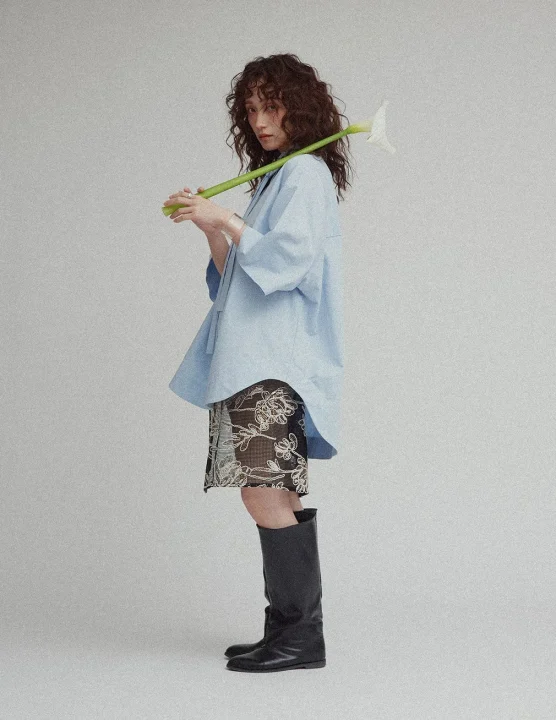 水色のオーバーサイズシャツと柄スカート、黒いロングブーツを着用し、白いカラーリリーを肩に抱えて立つ女性