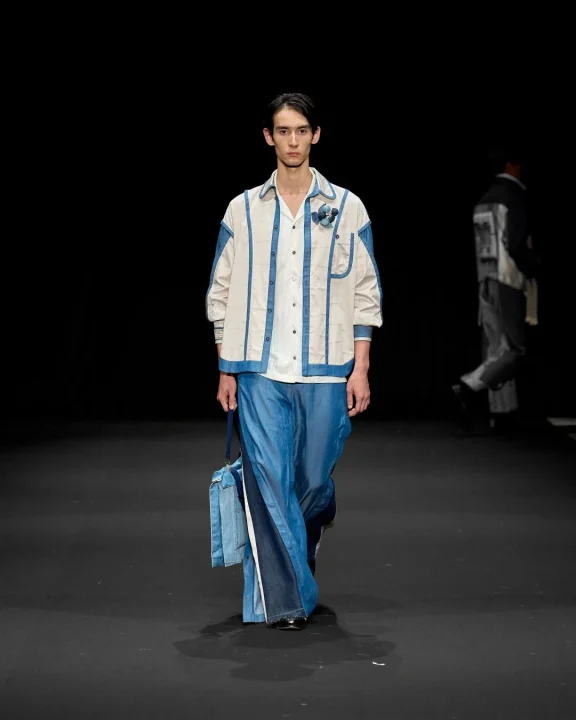 暗いランウェイを歩く男性モデルが、デニムと白の生地を組み合わせたユニークなレイヤード衣装と、それに合わせた青いバッグを披露しています。