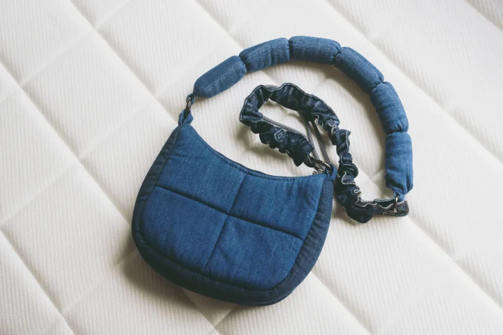 白いキルティングの背景に置かれた、デニム生地のキルティング加工されたショルダーバッグです。カジュアルなデザインで、普段使いにぴったりのアイテムです。