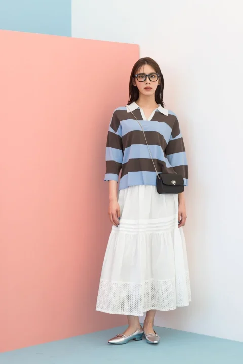 ピンクと水色の壁を背景に、ボーダーのラガーシャツと白いロングスカートを着用し、眼鏡をかけた女性がポーズをとっている。カジュアルで爽やかな夏のコーディネート。