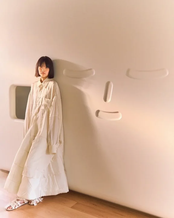 白いロングワンピースを着たショートヘアの女性が、抽象的な顔のレリーフがある白い壁にもたれかかっている。ミニマルで洗練された雰囲気のファッションポートレート。