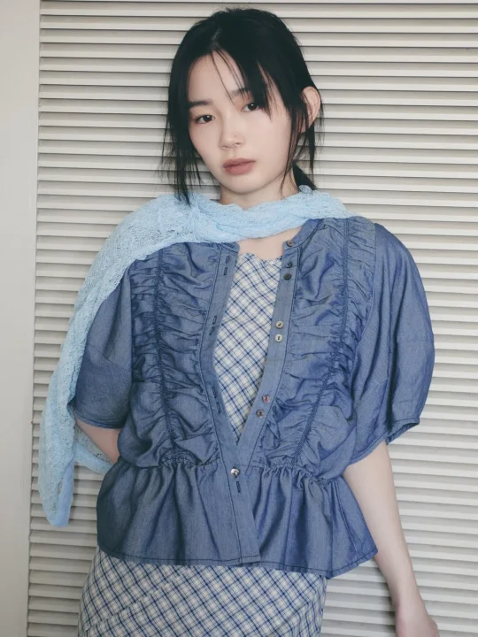 白いルーバーを背景に、青いデニム風のトップスとチェック柄のインナーを着た若い女性