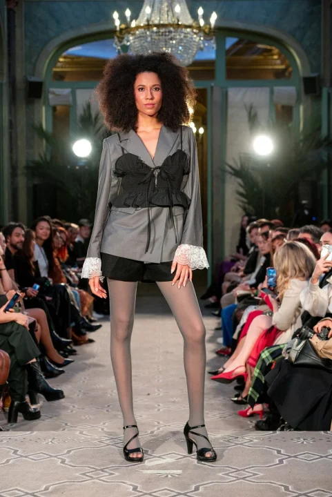 アフロヘアのモデルがグレーのブレザーと黒のビスチェ、ショートパンツ、グレーのタイツを着用してランウェイを歩いている