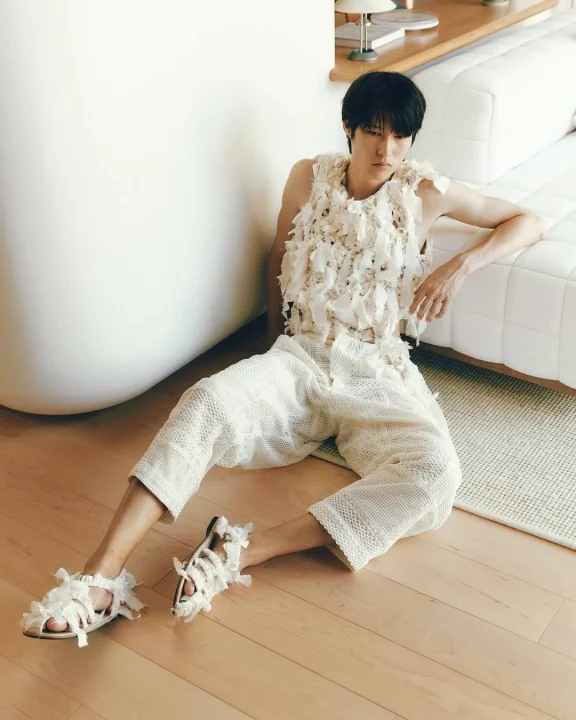 白いテクスチャーのある服とニットのパンツを着用した男性が、木製の床に座り、白い壁とソファを背景にしています。独特なサンダルも特徴的で、全体的にモダンでミニマリストな雰囲気が漂うファッションポートレートです。