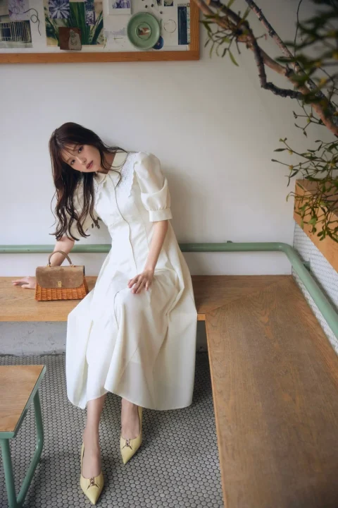 白いレース襟のワンピースを着た女性が、カフェのような明るい空間で木製ベンチに座っています。カゴバッグと黄色いパンプスがアクセントのエレガントでナチュラルなファッションポートレートです。