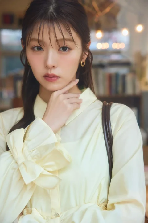 淡いイエローのブラウスを着た若い女性のポートレート。顎に手を添え、カメラをじっと見つめる表情が印象的です。背景はぼかされており、カフェのような落ち着いた室内で、自然体ながらも洗練された雰囲気が漂っています。