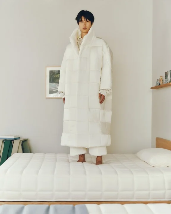 白いキルティングのオーバーサイズコートを着た男性が、白いマットレスの上に立っています。シンプルな白い壁の部屋で、ミニマリストな雰囲気が漂っています。