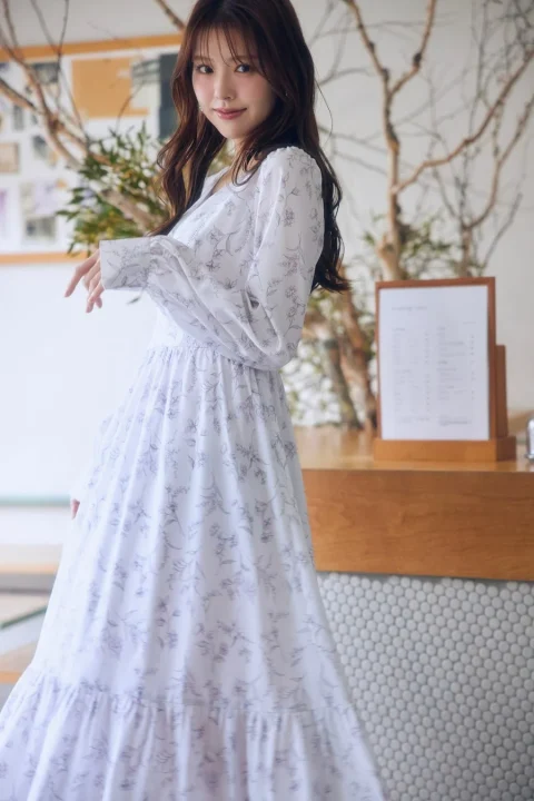 白い花柄のロングワンピースを着た女性が、カフェのような明るい空間で優しく微笑んでいます。長い髪が印象的で、自然光が差し込む中でリラックスした雰囲気です。