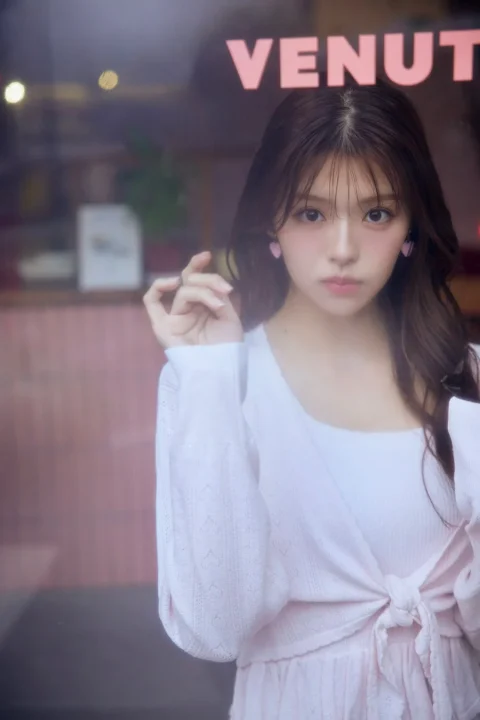 窓越しに撮影された若い女性のポートレート。ピンクのハート型イヤリングと白いトップス、ピンクのカーディガンを着用し、カメラをまっすぐに見つめている。柔らかい光に包まれ、可愛らしい雰囲気が漂う一枚です。