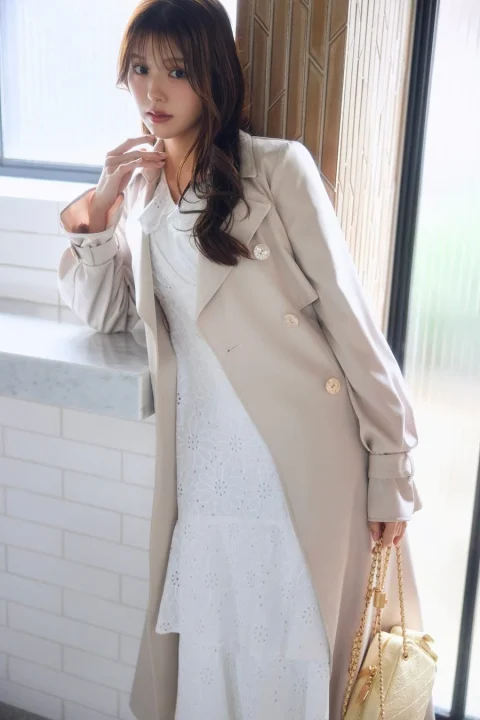 ベージュのトレンチコートと白い花柄刺繍ワンピースを着用した女性が、窓辺でポーズをとっています。上品でフェミニンな雰囲気のポートレートです。