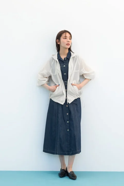 白い壁を背景に立つ女性の全身ショット。デニムのロングワンピースに、透け感のある白いフード付きパーカーを羽織り、両手をポケットに入れています。カジュアルで抜け感のある春夏のファッションコーディネートです。