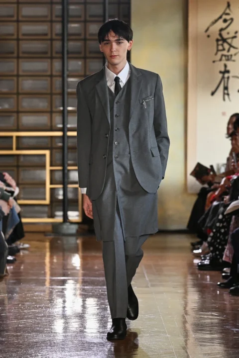 独特なデザインのグレーのスリーピーススーツを着用した男性モデルがランウェイを歩いている。