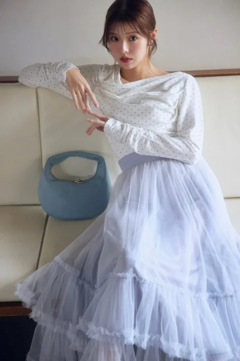 白いドット柄のトップスと淡いブルーのチュールスカートを着用した女性が、ソファに座ってポーズをとっています。隣には同色のハンドバッグが置かれ、上品でフェミニンな雰囲気が漂うポートレートです。