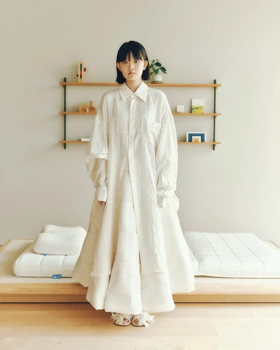 白いレイヤードワンピースを着た若い女性が、ミニマリストな空間に立つポートレート。ローベッドや木製の棚がある部屋で、穏やかで自然体なライフスタイルを感じさせます。