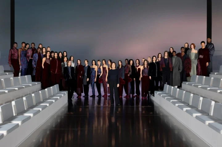 ファッションショーのランウェイで、多くのモデルとデザイナーが並んでポーズをとっている様子。