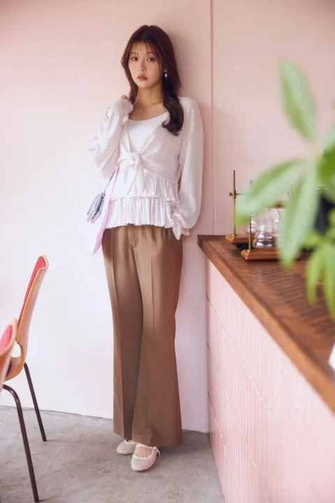 ピンクの壁を背景に、白いフリルのトップスとブラウンのワイドパンツ、きらめくバレエシューズを着用した若い女性が立っています。カフェのような空間で、落ち着いた雰囲気のファッションポートレートです。