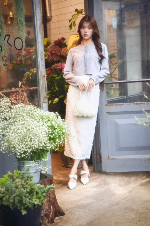 水色のカーディガンと白いレーススカートを着用した女性が、花屋の店頭に立つ全身ショット。白いビーズバッグを持ち、エレガントな春のファッションを披露している。