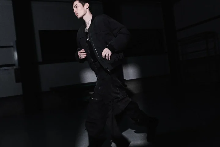 暗い室内を歩く若い男性の全身像です。黒いジャケットとパンツを着用し、スポットライトのような照明が当たっています。クールでミステリアスな雰囲気を醸し出しています。