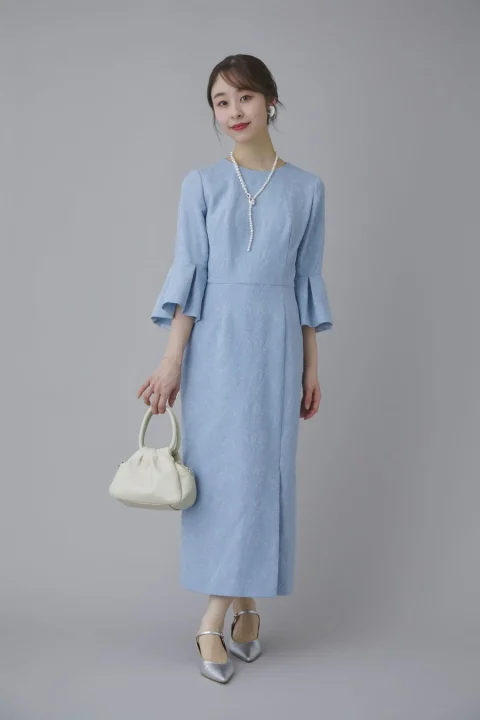 水色のエレガントなワンピースを着た女性が、パールネックレスと白いハンドバッグを身につけ、笑顔でポーズをとっています。