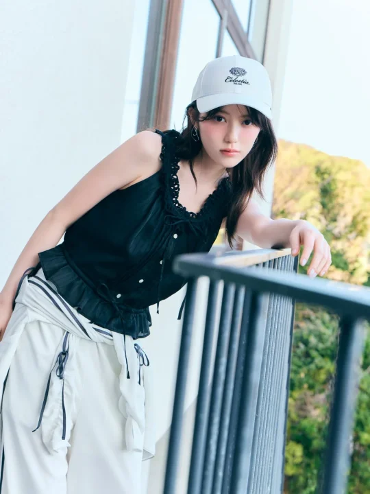 白いキャップと黒いノースリーブ、白いパンツ姿の若い女性がバルコニーの手すりに寄りかかっています