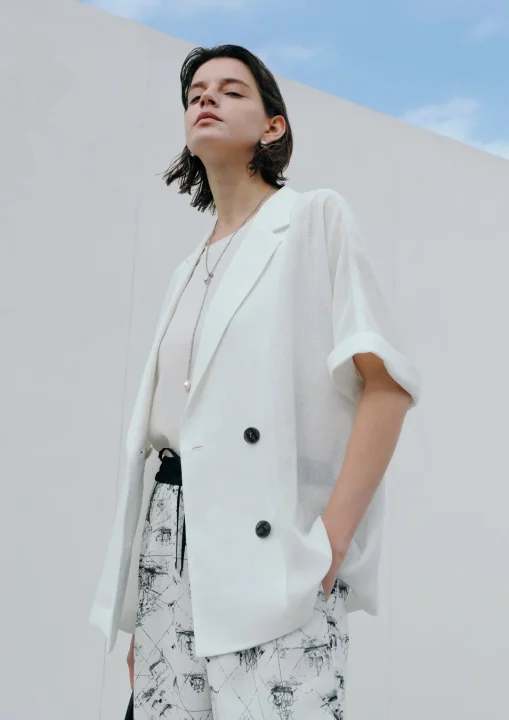 白いオーバーサイズジャケットと柄物のパンツを着用した女性のポートレート。屋外の白い壁と青空を背景に、洗練されたカジュアルなスタイル。