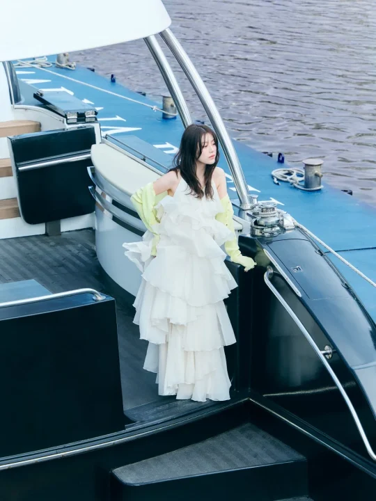 白いフリルのドレスと鮮やかなグローブを身につけた女性がヨットのデッキに立つポートレート