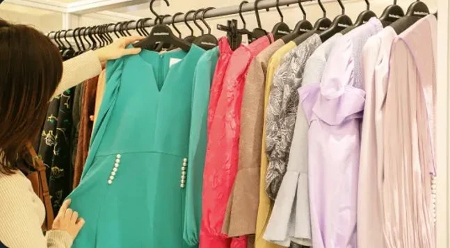 女性が洋服店で様々な色のドレスが並んだハンガーラックから、ターコイズブルーのドレスを選んでいる様子。