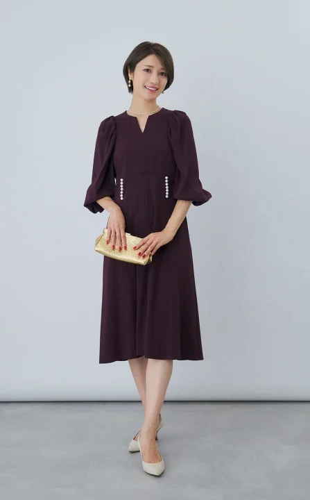 笑顔の女性が、Vネックの紫色の七分袖ワンピースを着用し、パールアクセサリーとゴールドのクラッチバッグを合わせた上品なフォーマルスタイルで立っている全身写真です。