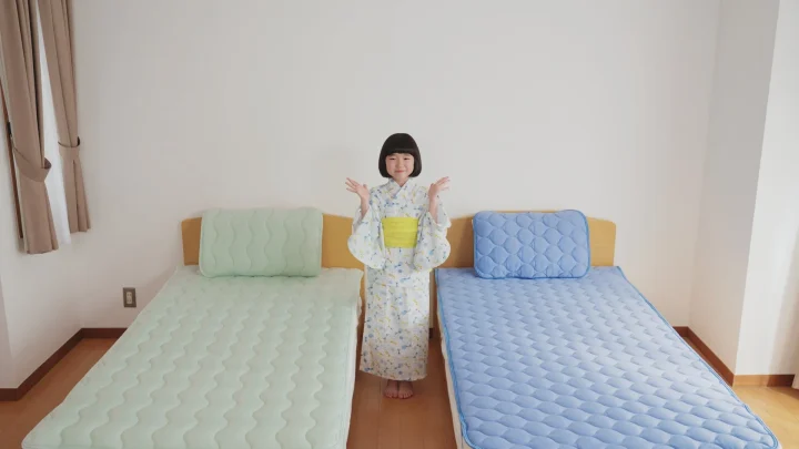 明るい部屋で、浴衣を着た日本人の女の子が2つのベッドの間に立って笑顔で両手を広げています。ベッドはそれぞれ緑と青の寝具で整えられています。
