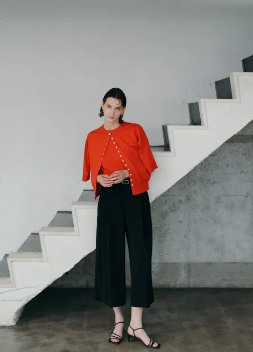 赤いカーディガンと黒いワイドパンツを着用したモデルが、白い階段のあるシンプルな空間に立っている。モダンでミニマルなファッションスタイル。
