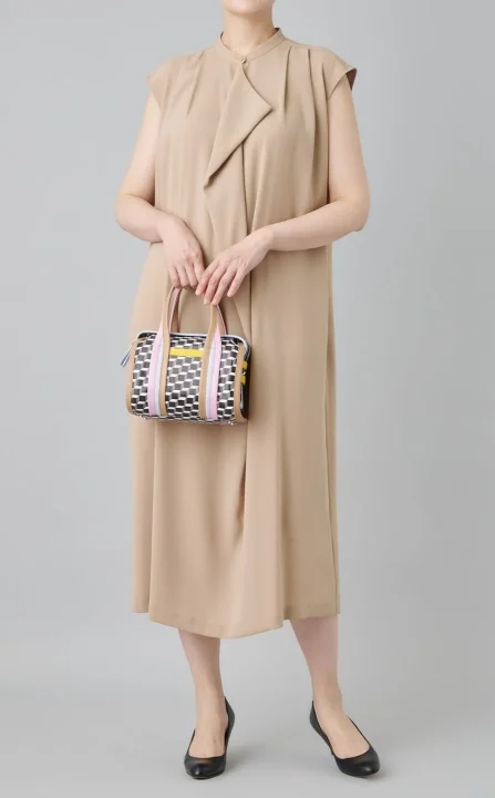 ベージュのノースリーブワンピースを着た女性が、幾何学模様とカラフルなストライプが特徴のハンドバッグを持ち、黒のフラットシューズを履いて立っています。