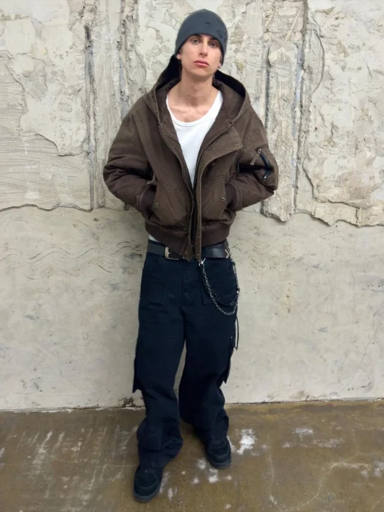 質感のある壁を背景に、ニット帽とブラウンのボンバージャケット、黒いカーゴパンツを着用した男性が立っています。ポケットに手を入れたカジュアルな立ち姿で、全身が写っています。