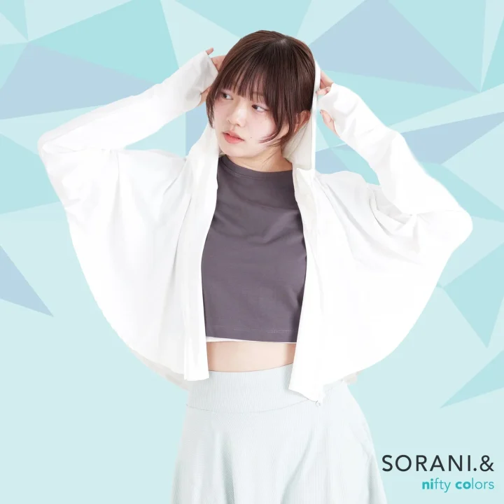 水色の幾何学模様の背景の前で、白いショート丈のパーカーを羽織り、フードに手をかけた女性のポートレートです。